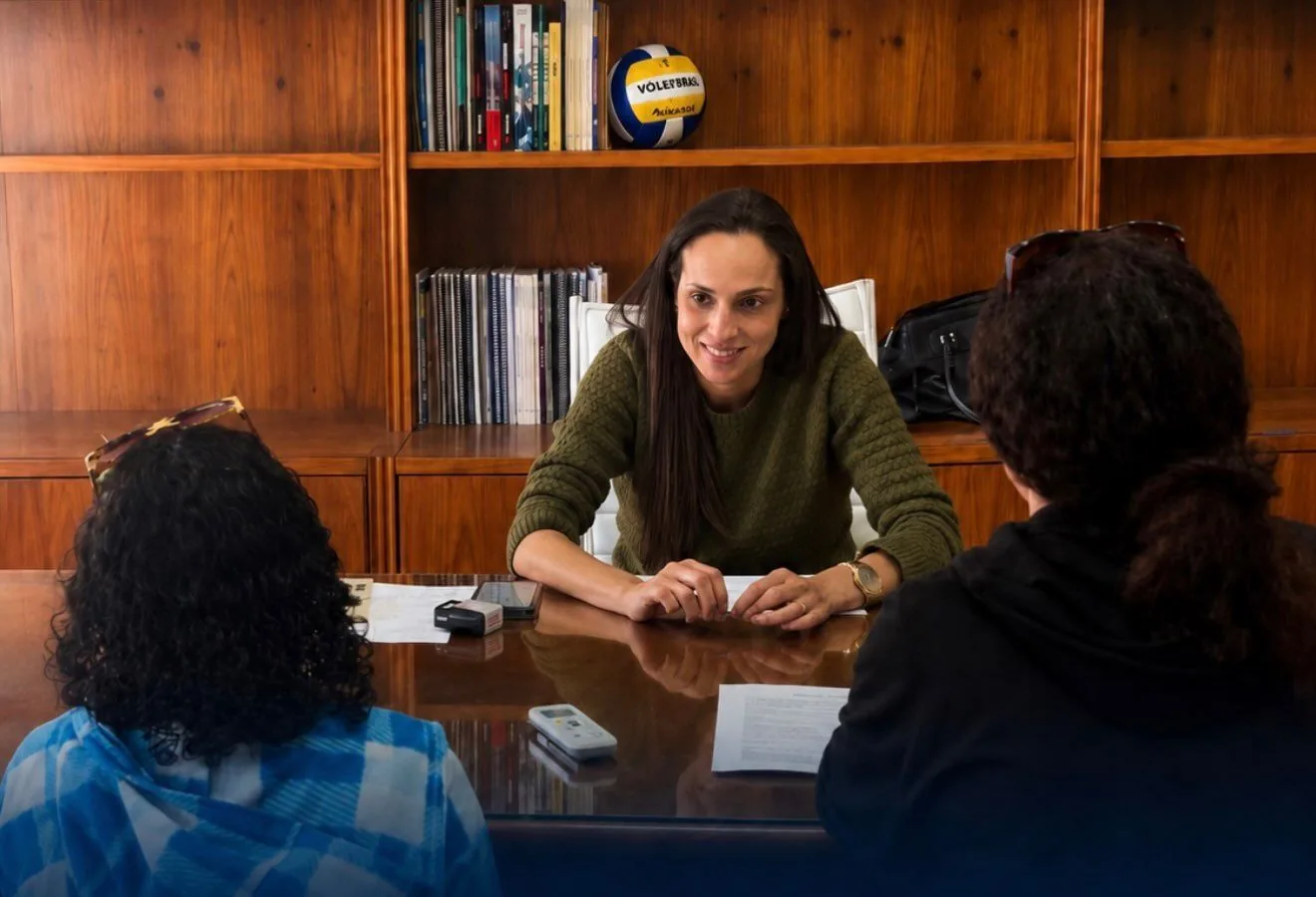 Paula do Vôlei em atendimento, sentada à mesa conversando com duas pessoas.