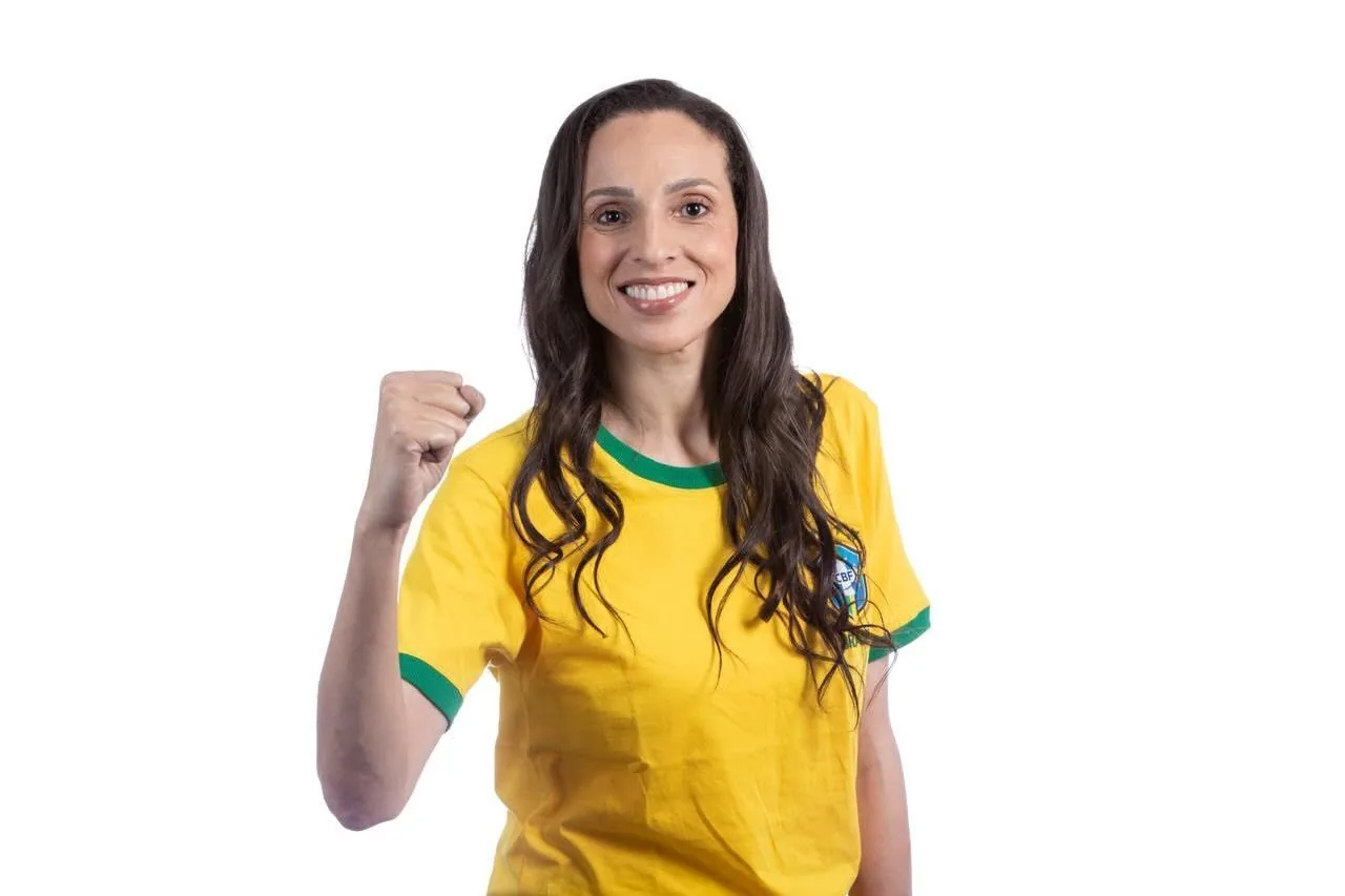 Paula do Vôlei sorrindo, de camisa amarela do Brasil, com o punho erguido em comemoração.