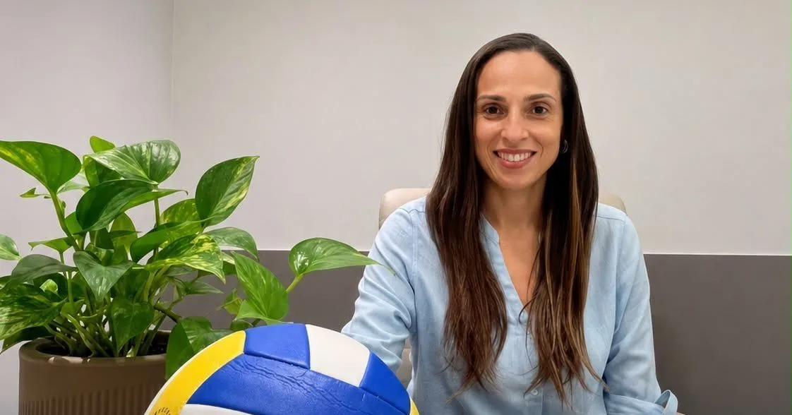 Paula do Vôlei sentada à mesa com uma bola de vôlei e medalhas de Superliga à sua frente.